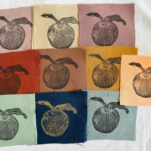 May include: Nine fabric squares with a black and white illustration of an apple on each. The squares are in various colors, including pink, beige, brown, orange, green, and blue.