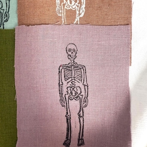 May include: Close-up of fabric swatches in shades of green, brown, and purple. Each swatch features a black and white illustration of a human skeleton. The skeletons are in a standing position, with detailed bone structures.