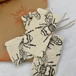 Könnte beinhalten: Drei handgefertigte Geschenkanhänger aus Recyclingpapier mit einem schwarzen Bienenmotiv. Die Anhänger sind mit einer Schnur gebunden.