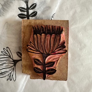 May include: A wooden printing block with a carved design of a flower with a pink and brown color scheme. The flower has a large, round head with many petals and a stem with leaves.
