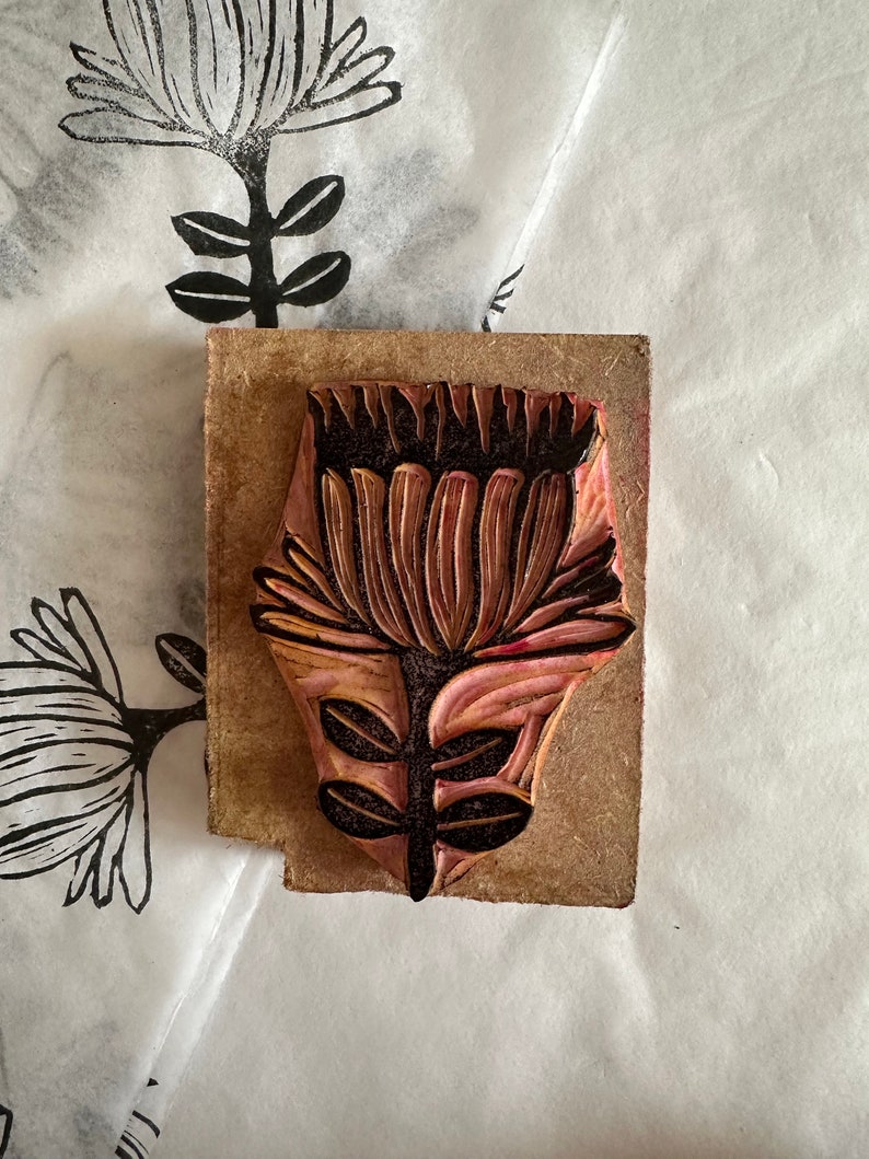 May include: A wooden printing block with a carved design of a flower with a pink and brown color scheme. The flower has a large, round head with many petals and a stem with leaves.