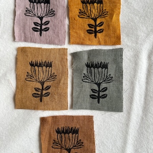 Könnte beinhalten: Fünf Stoffquadrate mit einem schwarzen, gedruckten Blumendesign. Die Quadrate sind in verschiedenen Farben: hellbraun, dunkelbraun, senfgelb, grau und helllila.