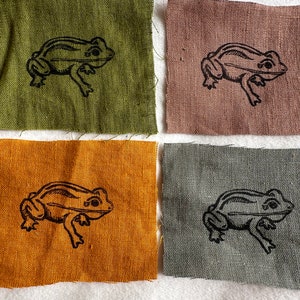 Pode incluir: Quatro quadrados de tecido com um desenho de sapo preto impresso em cada um. Os quadrados são de cores diferentes: verde, marrom, laranja e cinza.
