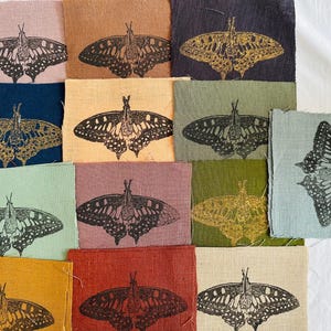 Peut inclure: Une collection de 12 carrés de tissu avec un motif de papillon noir imprimé sur chacun. Les carrés de tissu sont de différentes couleurs, notamment le rose, le marron, le bleu, le vert et le rouge.