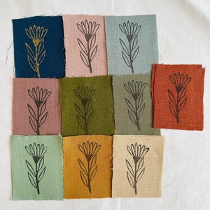 May include: Nine fabric squares featuring a black floral print in the centre. The squares are in various colours, including teal, pink, light blue, green, brown, beige, light green, orange, and natural.