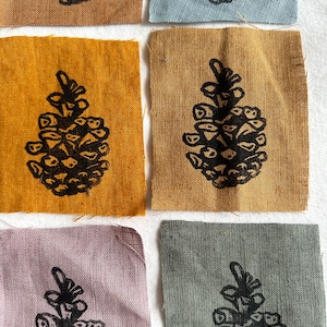 May include: Six fabric squares with black pinecone prints in shades of gold, tan, blue, grey, and lavender. The fabric has a textured appearance. The image is a close-up of the squares.