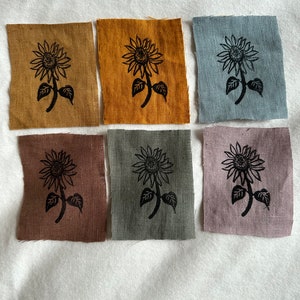Könnte beinhalten: Sechs Stoffquadrate mit einem schwarzen Sonnenblumen-Druck. Die Quadrate sind in verschiedenen Farben, darunter Senfgelb, Braun, Grau und Hellblau.