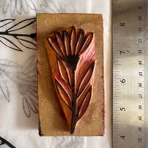 Puede incluir: Un bloque de impresión de madera con un diseño tallado de una flor rosa con un tallo y hojas marrones oscuros.