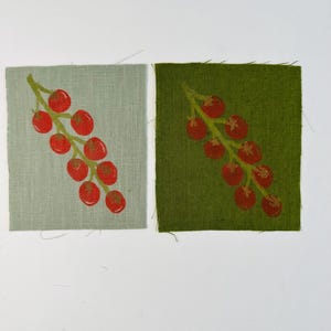 May include: Two fabric squares, one grey and one green, each feature a print of a red cherry branch. The cherries are a vibrant red, and the stems are green. The fabric has a textured appearance.