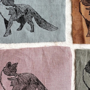 以下が含まれることがあります： 繰り返しのキツネのデザインが施された4つの布の正方形のクローズアップ。キツネは黒色で、生地は青、ピンク、オレンジ、緑の色合いです。キツネは詳細で様式化されたイラストで描かれています。