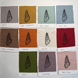 Op de afbeelding: Een verzameling stoffen stalen in verschillende kleuren, elk met een zwart schelpmotief. Kleuren zijn grijs, licht kaneel, mintgroen, mintblauw, echt kaneel, oranje, wit, rood, olijf, lavendel en zachtroze. Elk staal heeft een label met zijn kleurnaam.