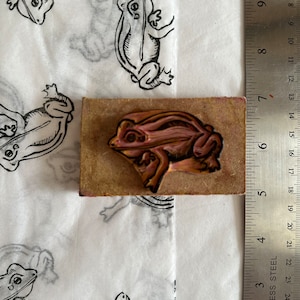 May include: A wooden frog stamp with a pink and brown frog design. The stamp is on a white surface with a ruler next to it.