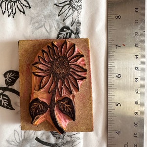 May include: A wooden printing block with a carved design of a sunflower with leaves. The block is approximately 2 inches wide and 3 inches tall. The sunflower is carved in a light pink color with a dark brown center. The leaves are carved in a dark green color.
