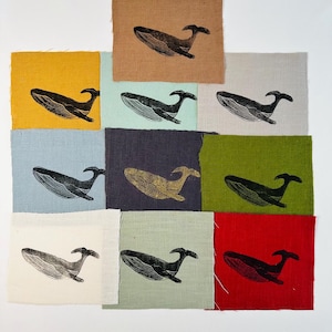 May include: A selection of fabric squares in diverse colours, each displaying a black whale illustration. The squares are arranged in a grid, showcasing the whale design on various backgrounds, including mustard, beige, and crimson.