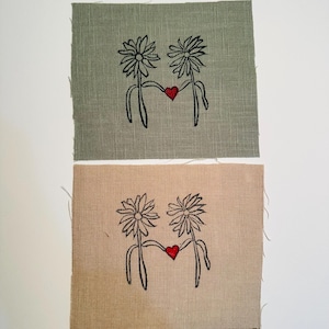 Op de afbeelding: Twee geborduurde vierkanten van stof met een ontwerp van twee gestileerde bloemen die een rood hart tussen hen vasthouden. Het bovenste vierkant is lichtgroen en het onderste vierkant is lichtbruin.