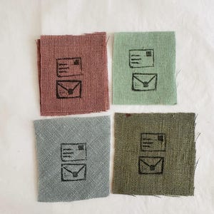 Peut inclure: Quatre petits carrés de tissu dans les tons rose poudré, vert sauge, bleu-gris et vert olive. Chaque pièce présente un tampon noir représentant une carte postale et une enveloppe. Les pièces sont disposées sur une surface blanche.