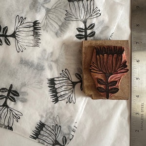 May include: A close-up of a wooden printing block with a detailed floral design. The block is used to create a print of a flower with a red center and black outline. The block is sitting on a white sheet of paper with several black and white prints of the same flower.