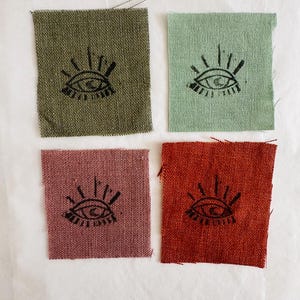 Può includere: Quattro quadrati di tessuto con un disegno di occhio nero stampato su ciascuno. I quadrati sono di colore verde, verde chiaro, rosa e rosso.