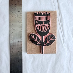 May include: A floral rubber stamp with a black flower design and pink details, mounted on a rectangular wooden block. A stainless steel ruler is placed next to the stamp for scale.