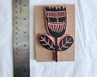 May include: A floral rubber stamp with a black flower design and pink details, mounted on a rectangular wooden block. A stainless steel ruler is placed next to the stamp for scale.