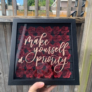 May include: A framed wall art piece with a black frame. The artwork features a background of red paper roses and the text "Make yourself a priority" in a cursive font. The piece is displayed against a wooden fence.