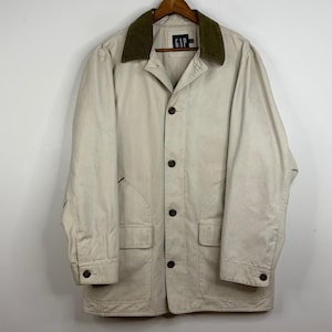 May include: A beige GAP jacket with a dark green corduroy collar. The jacket features a button-up front, two front pockets, and the GAP logo on the collar. It is suspended from a wooden hanger.