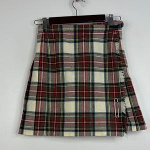 May include: A tartan skirt featuring a cream base with red, dark green, and thin blue stripes. The skirt has a wrap-style front with a fringed edge, a black leather side strap with a buckle, and a large silver-tone metal safety pin closure.