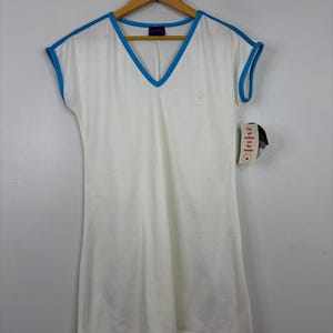 May include: A white V-neck dress with bright blue trim along the neckline and sleeve edges. The dress features a small embroidered logo on the chest and a hanging tag that reads, I'm all cotton on the inside, with a small red maple leaf icon.