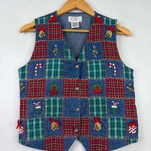 Peut inclure: Un gilet sans manches présentant un motif patchwork composé de carrés en jean bleu et de tissu à carreaux rouge et vert. Le gilet est orné de motifs de Noël brodés incluant des sapins, des sucres d'orge, des couronnes, des cloches et des boules.