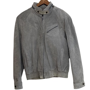 May include: A grey suede leather jacket with a zip closure and a stand-up collar. The jacket has two pockets on the front and a zipped pocket on the left sleeve.