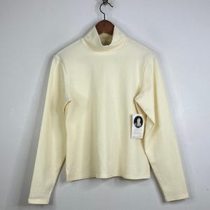 May include: Cream-coloured turtleneck long-sleeve shirt hanging on a wooden hanger. The shirt has a high neckline and a tag attached to the front. The fabric appears to be a soft, knit material. The shirt is a solid colour.