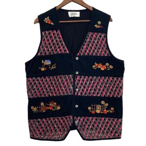 May include: A black quilted vest with a plaid pattern and embroidered autumn-themed designs. The vest has a V-neck and button closure. The embroidery includes a house, pumpkins, sunflowers, and a wheelbarrow.