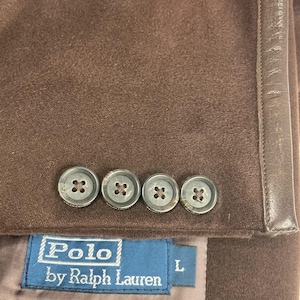 May include: A close-up of a brown blazer with four grey buttons. The blazer features a label reading "Polo by Ralph Lauren" in white text on a blue background. The size is "L".