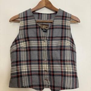May include: A grey plaid waistcoat with a V-neck and button-up front. The waistcoat features a pattern of grey, black, red, and white lines. It is hanging on a wooden hanger.