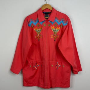 May include: A coral-coloured jacket with a zip-up front and long sleeves. The jacket features colourful embroidered geometric patterns in turquoise, yellow, orange, and black. The collar has a label that reads "18 Collection".