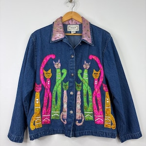 Peut inclure: Une veste en jean bleu ornée d'un motif appliqué coloré représentant six chats stylisés dans des tons rose, vert, jaune et violet. La veste présente une fermeture boutonnée et un col lavande à motifs. L'étiquette indique Tantrums.