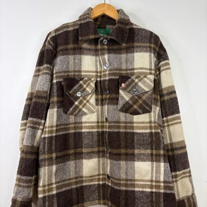 Peut inclure: Une chemise à manches longues boutonnée avec un motif à carreaux marron, crème et beige. La chemise comprend un col classique, une fermeture boutonnée sur le devant et deux poches poitrine à rabats boutonnés. Le tissu a un aspect texturé.