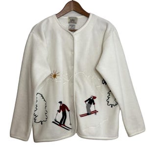 May include: Cream-coloured fleece jacket with a button-up front and long sleeves. The jacket features embroidered mountain and skier designs in red, black, and brown. A sun design is also embroidered on the upper left side. The jacket is hanging on a wooden hanger.