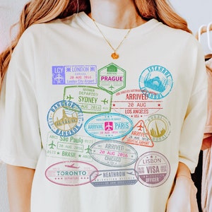 以下が含まれることがあります： カラフルなパスポートスタンプのグラフィックプリントが施された薄い黄色のTシャツ。スタンプには、ロンドン、プラハ、シドニー、ロサンゼルス、パリ、トロント、リスボンなど、さまざまな都市や国が描かれています。スタンプのテキストには、到着日と出発日、都市名、空港コードが含まれています。