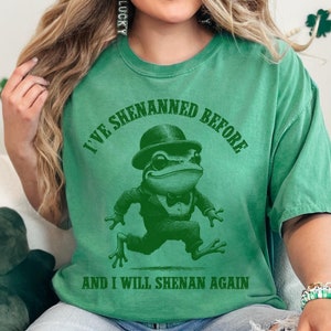 Puede incluir: Una camiseta verde con un gráfico de una rana con un sombrero de copa y una pajarita. El texto en la camiseta dice "I've Shennaned Before and I Will Shenan Again".