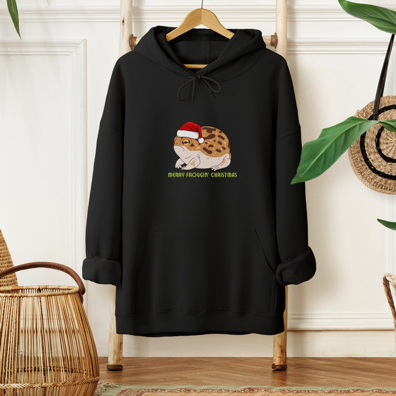 Op de afbeelding: Zwart hoodie met een afbeelding van een kikker met een kerstmanmuts. De tekst "Merry Froggin' Christmas" staat onder de kikker.