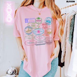 以下が含まれることがあります： ピンクのオーバーサイズTシャツに、カラフルなパスポートスタンプのグラフィックプリントが施されています。スタンプには、ロンドン、シドニー、パリ、東京などの都市名が記されています。スタンプのテキストには、到着日と出発日が含まれています。