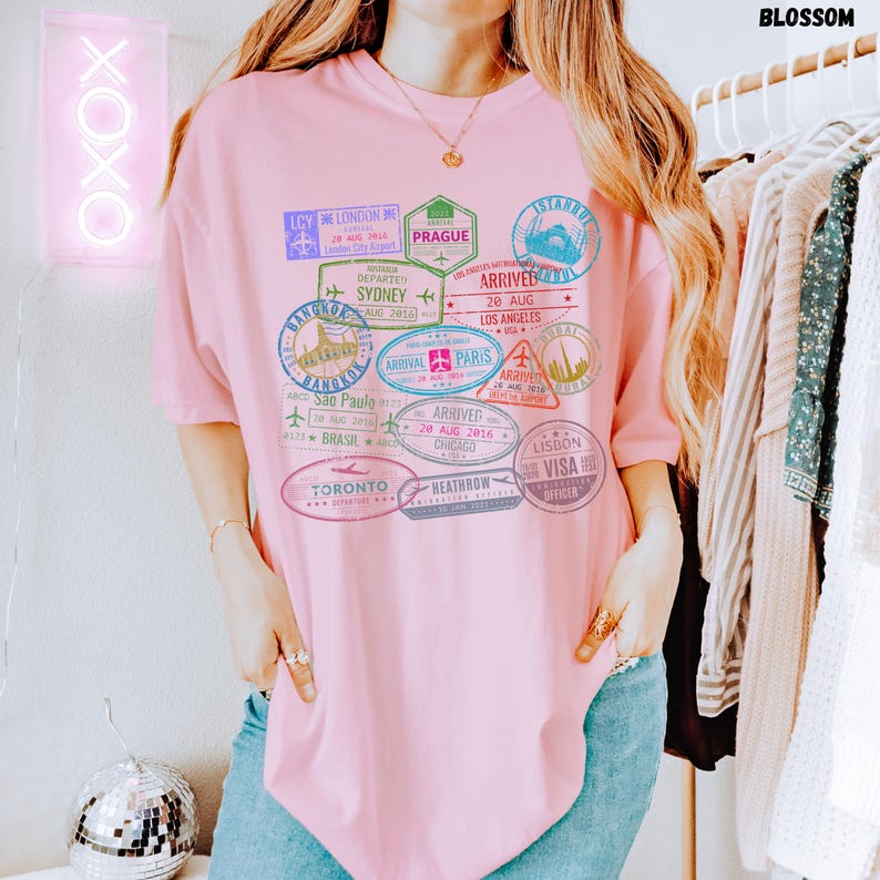 以下が含まれることがあります： ピンクのオーバーサイズTシャツに、カラフルなパスポートスタンプのグラフィックプリントが施されています。スタンプには、ロンドン、シドニー、パリ、東京などの都市名が記されています。スタンプのテキストには、到着日と出発日が含まれています。