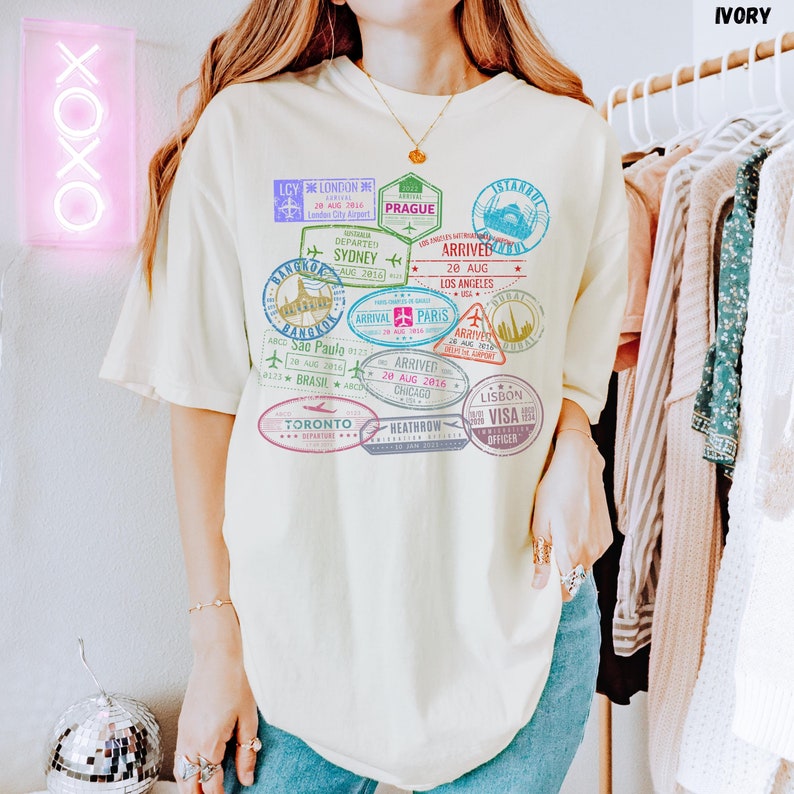 以下が含まれることがあります： カラフルなパスポートスタンプのグラフィックプリントが施された薄い黄色のTシャツ。スタンプには、ロンドン、プラハ、シドニー、ロサンゼルス、パリ、トロント、リスボンなど、さまざまな都市や国が描かれています。スタンプのテキストには、到着日と出発日、都市名、空港コードが含まれています。