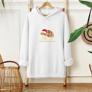 Op de afbeelding: Witte hoodie met een cartoon kikker die een kerstmanmuts draagt. De tekst "Merry Frogg'in Christmas" staat onder de kikker.