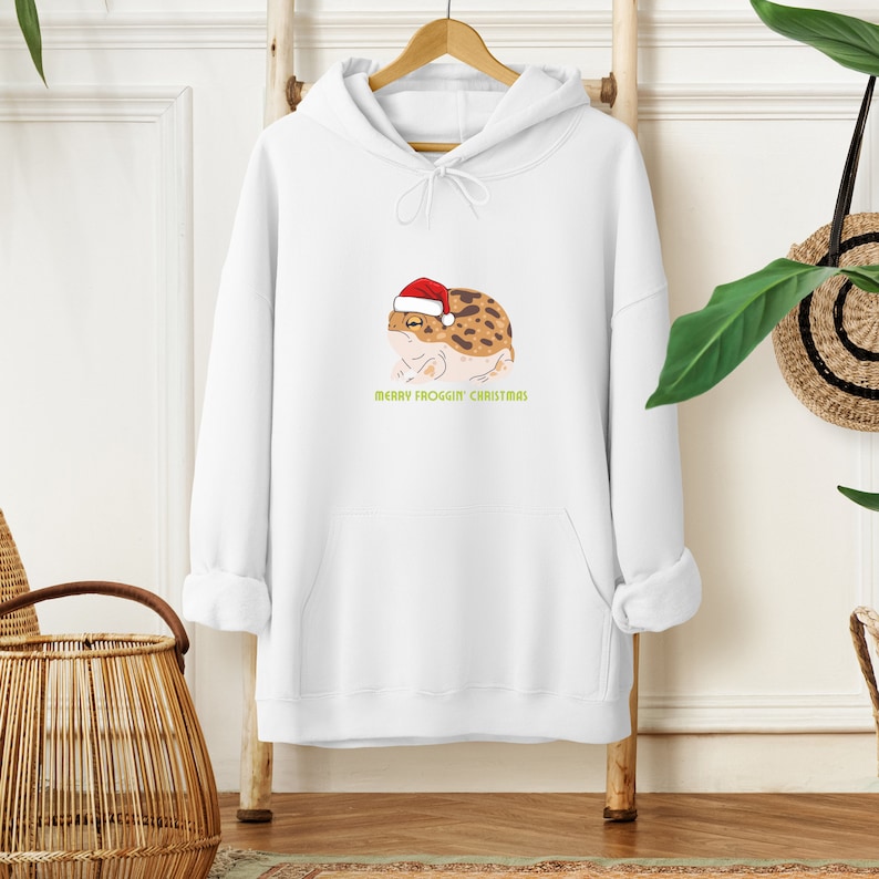 Op de afbeelding: Witte hoodie met een cartoon kikker die een kerstmanmuts draagt. De tekst "Merry Frogg'in Christmas" staat onder de kikker.
