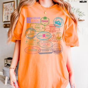 以下が含まれることがあります： オレンジ色のTシャツに、ロンドン、プラハ、イスタンブール、シドニー、ロサンゼルス、パリ、シカゴ、トロント、ヒースロー、リスボンなど、世界中のさまざまな都市のカラフルなパスポートスタンプのグラフィックプリントが施されています。