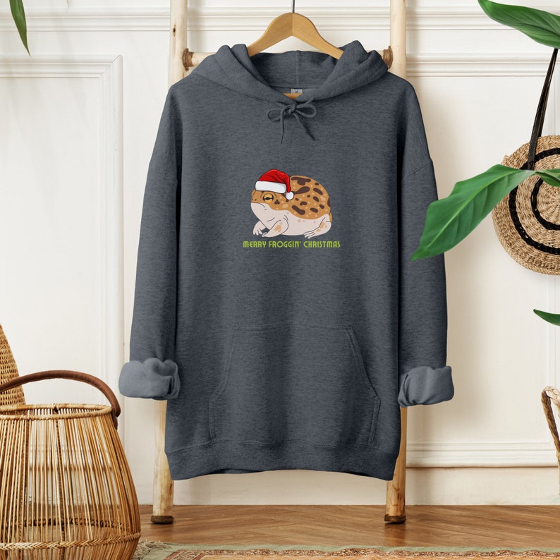 Op de afbeelding: Een donkergrijs hoodie met een afbeelding van een kikker die een kerstmanmuts draagt. De tekst "Merry Frogg'in Christmas" staat onder de kikker.