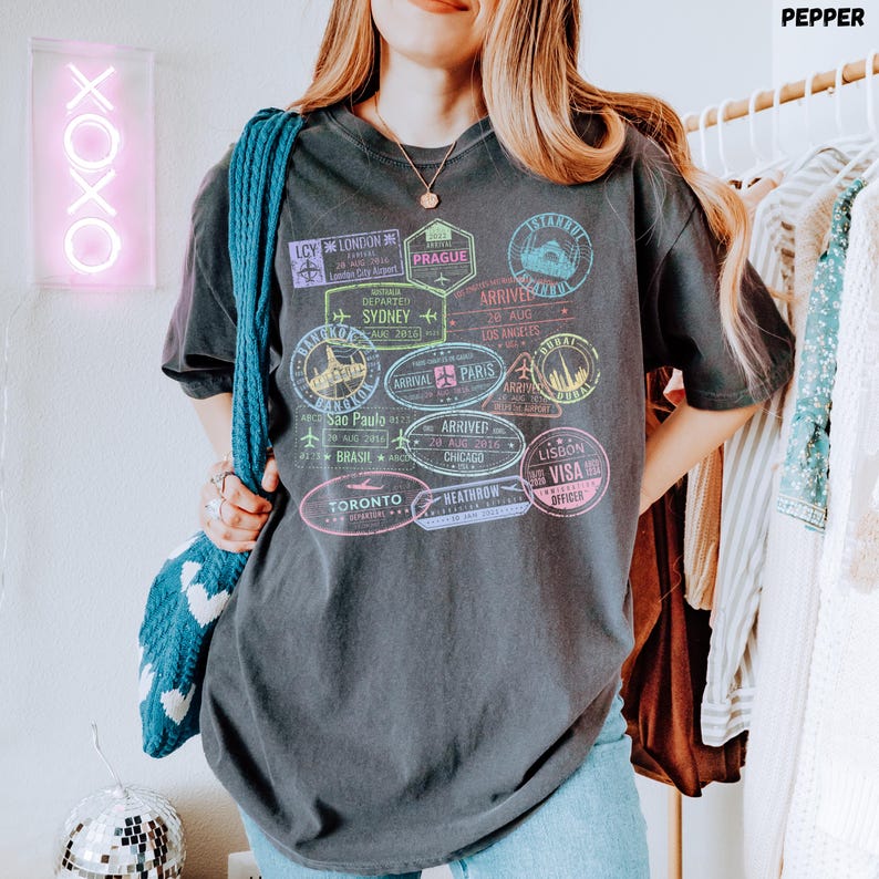 以下が含まれることがあります： カラフルな旅行スタンプのグラフィックプリントが施された黒いTシャツ。スタンプには、ロンドン、プラハ、シドニー、ロサンゼルス、パリ、シカゴ、トロント、リスボンなどの都市名が記載されています。スタンプのテキストには、「出発」、「到着」、「ビザ担当官」、「ヒースロー」などが含まれています。