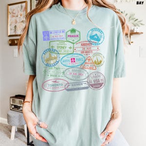 以下が含まれることがあります： 薄い青色のTシャツに、ロンドン、プラハ、イスタンブール、シドニー、バンコク、パリ、ドバイ、サンパウロ、シカゴ、リスボン、トロントなど、世界中のさまざまな都市のカラフルなパスポートスタンプのグラフィックプリントが施されています。スタンプには、到着日と出発日、都市名、空港コードが記載されています。